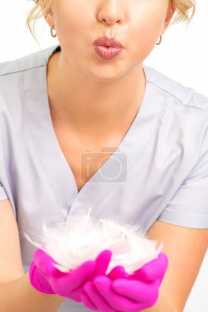 Foto de Joven hermosa mujer esteticista en guantes protectores de color rosa de pie y sosteniendo plumas blancas sobre fondo blanco de la pared - Imagen libre de derechos