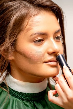 Téléchargez les photos : Belle jeune femme brune recevant du maquillage avec un correcteur de bâton sur son visage dans un salon de beauté - en image libre de droit