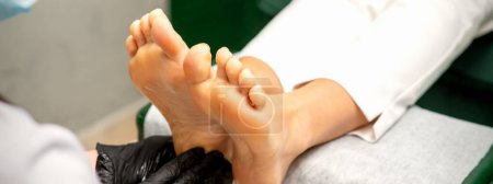 Téléchargez les photos : Pédicure hydratant pieds féminins avec lotion portant des gants de protection noirs dans un salon de beauté - en image libre de droit