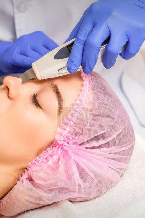 Téléchargez les photos : Jeune femme caucasienne recevant un nettoyage de la peau du visage par ultrasons équipement de cosmétologie visage dans un salon médical - en image libre de droit