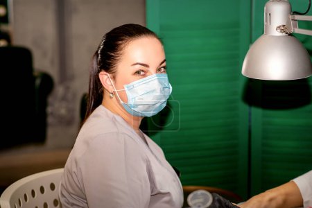 Téléchargez les photos : Jeune podiatre féminine avec un masque protecteur regardant la caméra dans sa clinique de podiatrie - en image libre de droit