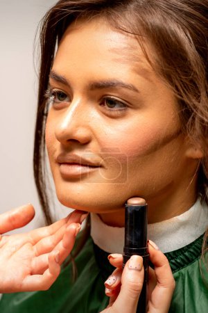 Téléchargez les photos : Belle jeune femme brune recevant du maquillage avec un correcteur de bâton sur son visage dans un salon de beauté - en image libre de droit