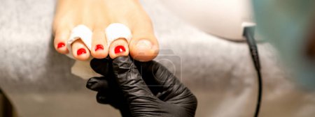 Foto de Proceso de pintura de uñas. Una mano maestra de manicura sostiene las uñas pintadas de rojo en una pierna femenina en un salón de belleza - Imagen libre de derechos