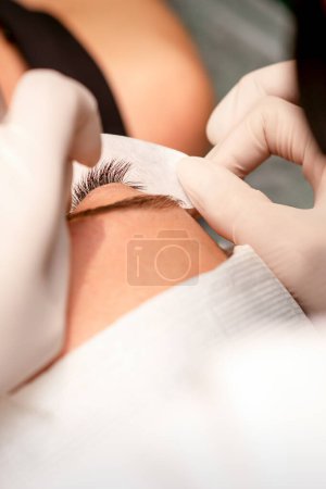 Téléchargez les photos : Les mains du cosmétologue collent du ruban blanc sous l'œil de la jeune femme caucasienne pendant la procédure d'extension des cils, gros plan - en image libre de droit