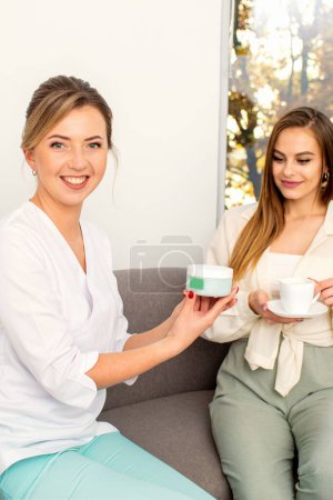 Téléchargez les photos : Esthéticienne offrant un produit pour la jeune femme tenant un pot en plastique blanc avec une crème assise sur le canapé - en image libre de droit