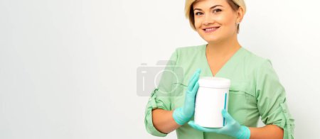 Photo for Cosmetics creams and skin care products in the hands of the female beautician smiling and standing over the white wall background - Royalty Free Image