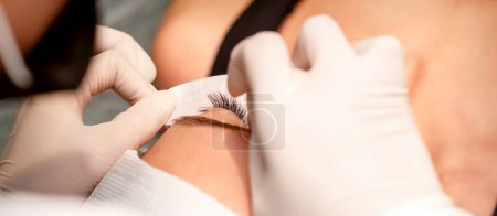 Photo for The hands of the cosmetologist are gluing white tape under the eye of the young caucasian woman during the eyelash extension procedure, closeup - Royalty Free Image