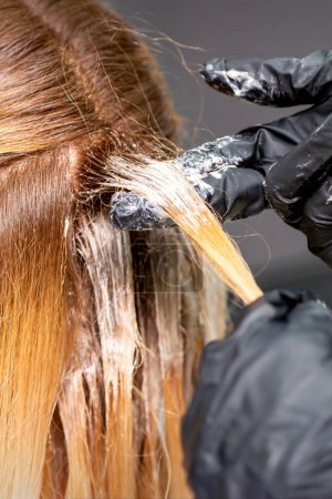 Photo for Hands of the hairdresser are applying the dye with gloved fingers on the red hair of a young red-haired woman in a hairdresser salon, close up - Royalty Free Image