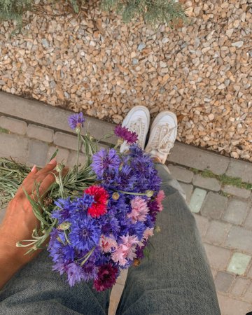 Photo for Photo a girl holds up a bouquet of blue, red and pink cornflowers against the background of legs and the ground - Royalty Free Image
