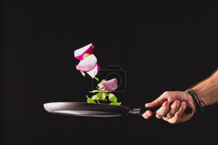 Foto de Hacer Ceviche al estilo brasileño sobre fondo negro. Freír la sartén arrojando ingredientes como cebolla roja, queso, albahaca y condimentos al aire. - Imagen libre de derechos