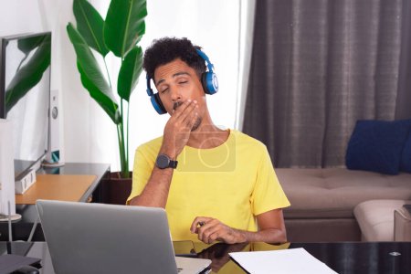Black Man Wearing Yellow T-shirt at Remote Job in His House. He is Tired at Desk With Laptop and Phones