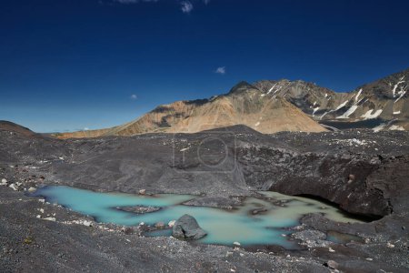 Turquoise glacial lake pool nestles among dark moraines under a stark mountain range and blue sky