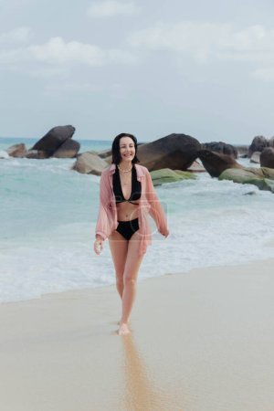Fröhliche Frau im Badeanzug spaziert an einem Strand, mit großen Felsen und dem Meer im Rücken
