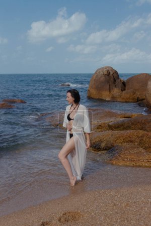 Woman in a white dress strolls beside the sea, with waves gently lapping at her feet and rocks in the background