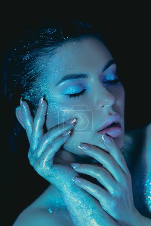Foto de Mujer con maquillaje brillante toca su cara, bañada en un brillo azul etéreo - Imagen libre de derechos