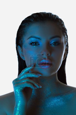 Woman with shimmering skin gently touches her face under a blue light, beauty makeup face