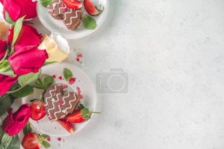  Valentinstag Dessert. Herzförmige Biskuitschokolade Mini-Kuchen mit Erdbeere und Minze. Draufsicht auf weißem Hintergrund mit Rosen Blumen Kopierraum