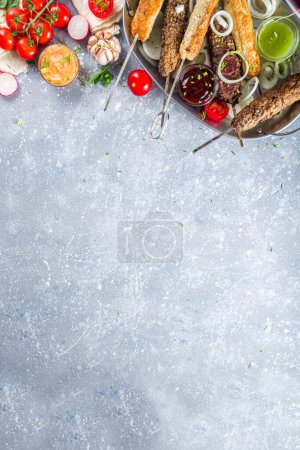 Photo for Summer bbq party. Shish kebab on a stick, from ground beef and chicken meat. Lula kebab, traditional Caucasian asian dish. With various sauces, spices and tomatoes, grey stone background, copy space above - Royalty Free Image