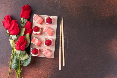 Foto de Sweet fruit sushi roll for Valentine day dating dinner or brunch. Mousse berry white chocolate creamy  sweet sushi dessert, with rose flowers bouquet on dark background top view copy space - Imagen libre de derechos