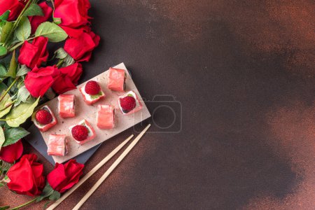 Foto de Sweet fruit sushi roll for Valentine day dating dinner or brunch. Mousse berry white chocolate creamy  sweet sushi dessert, with rose flowers bouquet on dark background top view copy space - Imagen libre de derechos