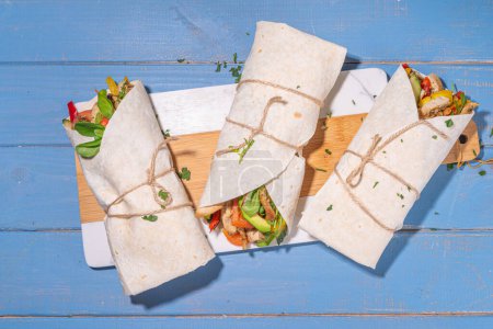 Classic street food shawarma or burrito, healthy sandwich wrapped in tortilla flat bread with fried chicken meat, fresh vegetables, sauce, on bright blue wood background, top view flat lay copy space