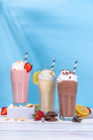Set of summer refreshing milkshakes or smoothies with classic delicious taste - vanilla lemon, strawberry, chocolate, with whipped cream and toppings, On a white wooden light blue sunny background 