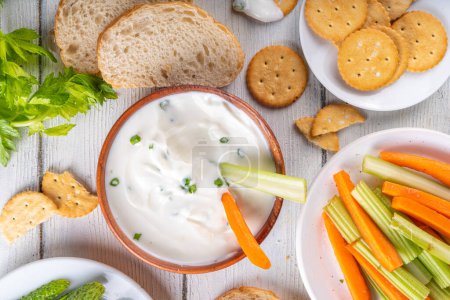 Hüttenkäse-Dip mit hohem Eiweißanteil mit frischem Gemüse, Brot und Crackern auf einem hölzernen weißen Tisch. Ernährung gesunder Quarkaufstrich mit verschiedenen Snacks