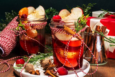 Photo for Hot mulled wine with cinnamon and star anise. Winter autumn fruit and berry grog, hot punch drink with orange, apple, cranberry, spices, with Christmas tree branches and gift boxes on wooden table - Royalty Free Image