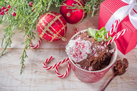 Téléchargez les photos : Mugcake à la menthe poivrée au chocolat. Petit déjeuner de Noël simple, recette d'idée de collation, gâteau au chocolat noir dans des tasses festives avec crème fouettée et morceaux de canne à bonbons garniture - en image libre de droit
