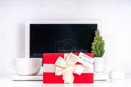 Photo for Present from secret santa on workplace table. Office Christmas and New Year celebration game. Office table surface, notebook with festive gift box, white background flatlay copy space - Royalty Free Image