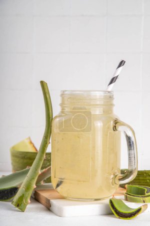Glass mug of fresh homemade aloe vera juice on white concrete table with fresh aloe leaves, copy space