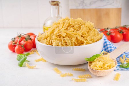 Raw uncooked Mafaldine pasta with ingredients for cooking. Short italian Mafaldine pasta with olive oil, cherry tomatoes, basil, spices on white kitchen table, background copy space