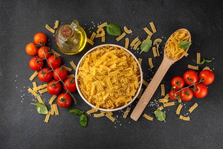 Raw uncooked Mafaldine pasta with ingredients for cooking. Short italian Mafaldine pasta with olive oil, cherry tomatoes, basil, spices on black kitchen table, background copy space