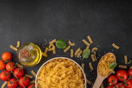 Raw uncooked Mafaldine pasta with ingredients for cooking. Short italian Mafaldine pasta with olive oil, cherry tomatoes, basil, spices on black kitchen table, background copy space