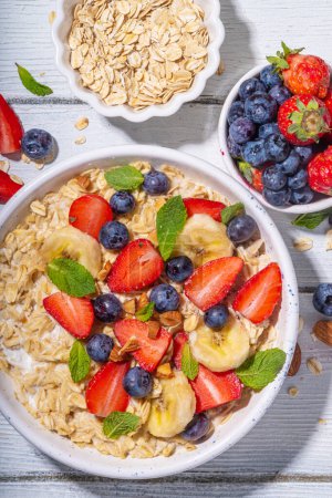 Schüssel süßes Frühstück Haferflocken mit Beeren vorhanden. Portionsteller Hafervollkornbrei mit Joghurt, frischen Bananen, Erdbeeren, Blaubeeren, Mandelnüssen, auf weißem Holztisch. Gesundes Sommerfrühstück