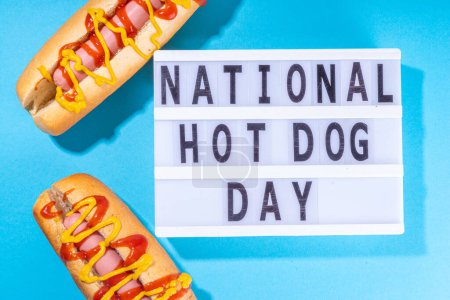 National Hot Dogs day background, hotdog summer party festival foods, Two tasty classic american hot dogs with sauces and lightbox sign with inscription National Hot Dog Day on blue background 