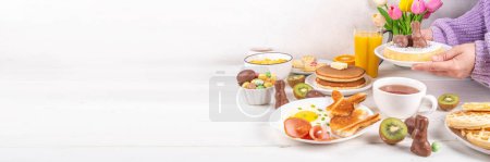 Foto de Vacaciones de Pascua desayuno dulce, almuerzo. Gofres belgas decorados con Pascua, panqueques, cereales para el desayuno y leche, huevos fritos con tostadas y tocino, huevos de chocolate de Pascua y conejo, con flores de primavera - Imagen libre de derechos