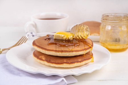 Stack of fresh baked homemade breakfast pancakes with butter and honey or maple syrup drizzles on white wooden table