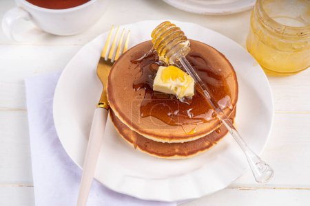 Stack of fresh baked homemade breakfast pancakes with butter and honey or maple syrup drizzles on white wooden table
