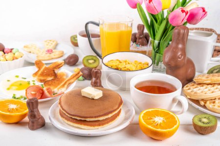 Easter holiday sweet breakfast, brunch. Easter decorated belgian waffles, pancakes, breakfast cereals and milk, fried eggs with toasts and bacon, Easter chocolate eggs and bunny, with spring flowers