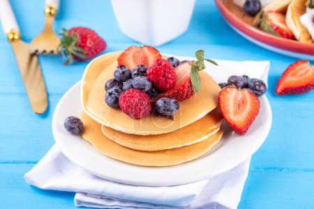 Stapel traditioneller amerikanischer Pfannkuchen mit Sommerbeeren. Frühstück süße Pfannkuchen mit Honig, Erdbeere und Blaubeere, am Morgen Sonne beleuchtet blauen Holzhintergrund Kopierraum