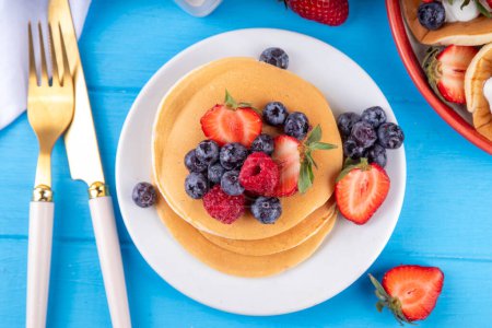 Stack of traditional american pancakes with summer berries. Breakfast sweet pancakes with honey, strawberry and blueberry, on morning sun lighted blue wooden background copy space