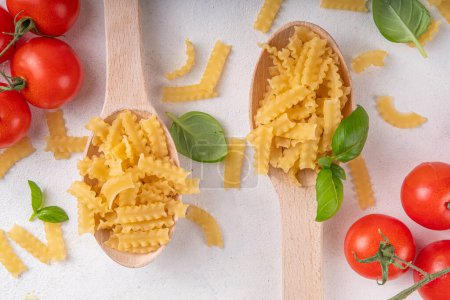 Raw uncooked Mafaldine pasta with ingredients for cooking. Short italian Mafaldine pasta with olive oil, cherry tomatoes, basil, spices on white kitchen table, background copy space