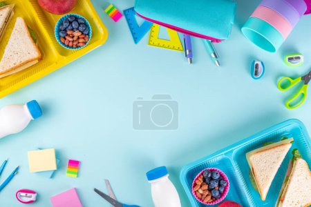 Healthy school meal, children packed lunch box with fruit, berry, nuts and sandwich with vegetables. Kids diet snack food with education school supplies, bright blue background top view copy space