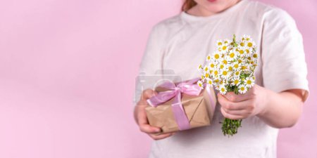 Photo for Birthday, Mother's Day international holiday greeting card. Child's hands holding a bouquet with wild flowers daisies and a gift box with a ribbon, on a pink background  copy space - Royalty Free Image