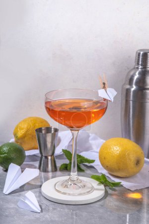 Photo for Paper plane boozy alcohol cocktail with Bourbon, whiskey, amaro, aperol aperitif, fresh lemon juice and paper plane craft made decor, on white background with hard light and bar utensils - Royalty Free Image