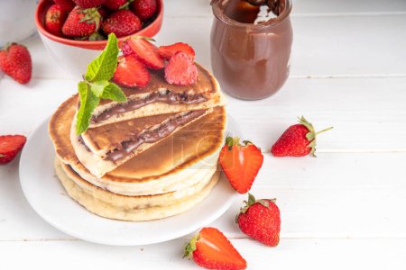 Stapelweise mit Schokolade gefüllte Nutella-Pfannkuchen. Süße Frühstückspfannkuchen mit Schokoladensauce und frischen Erdbeeren auf weißem Küchentisch