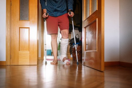 Foto de Hombre herido con el pie vendado caminando en casa con bastones. - Imagen libre de derechos