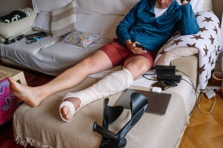 Photo for Injured man with bandaged foot using a smartphone. - Royalty Free Image