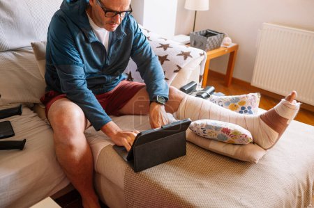 Téléchargez les photos : Homme blessé avec un pied bandé à l'aide d'une tablette. - en image libre de droit